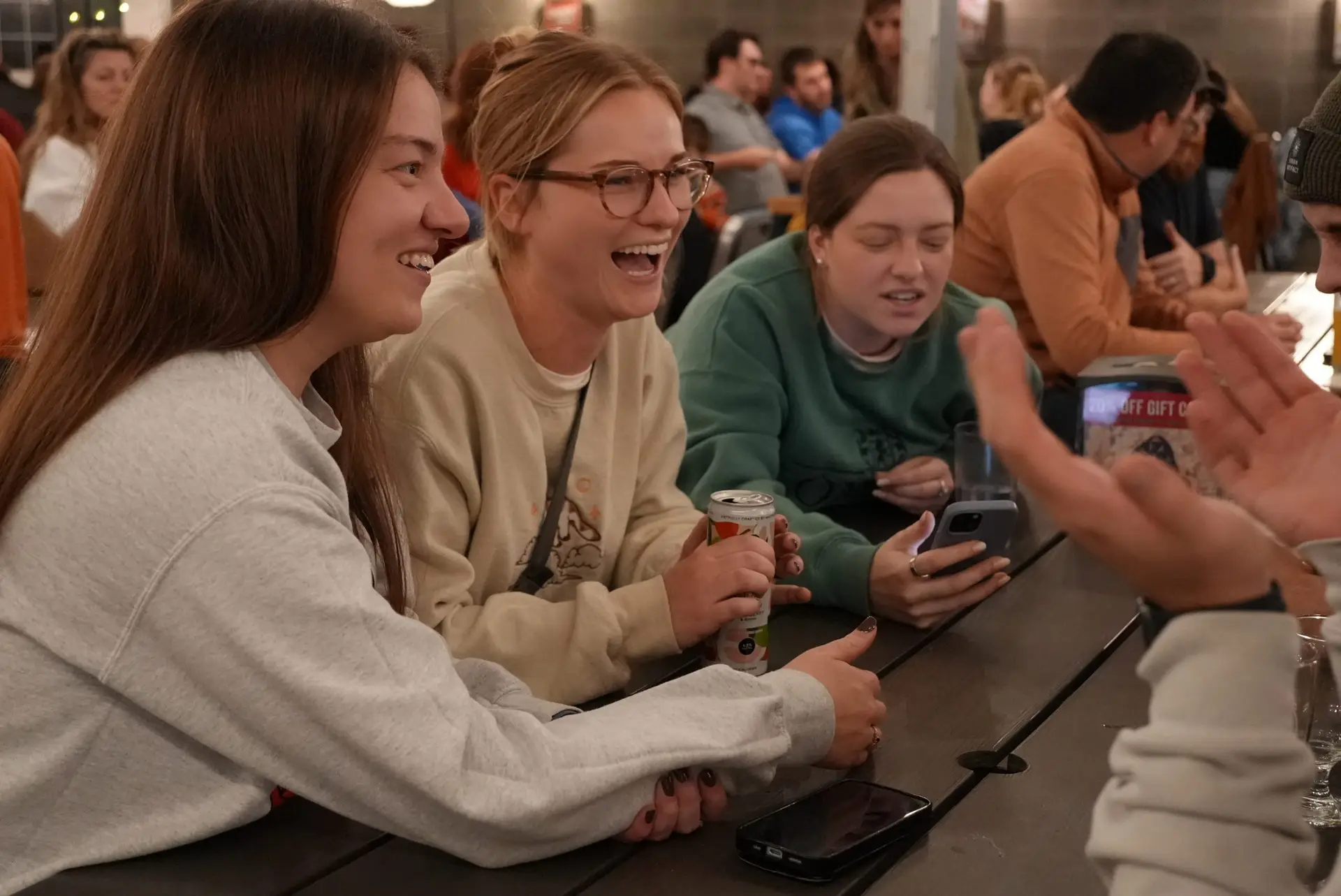 Three women laughing and holding drinks at a long bar table during trivia night