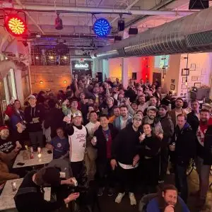 Large group of people smiling and raising drinks at a packed trivia night event in a bar