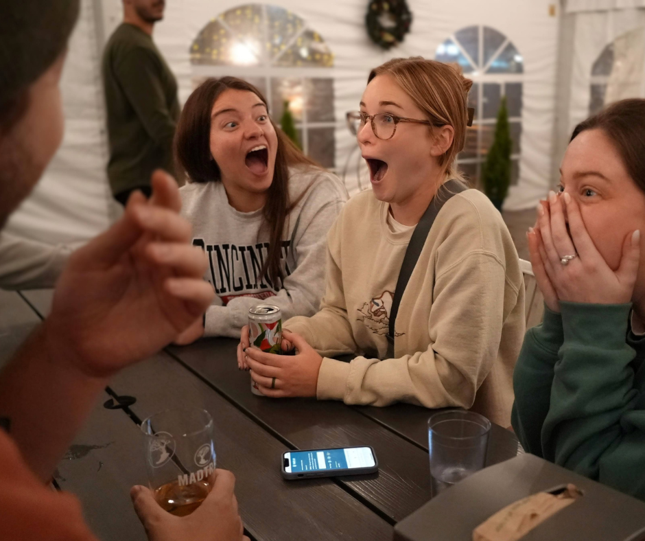 Three women reacting with excitement and surprise during a lively conversation at a bar table