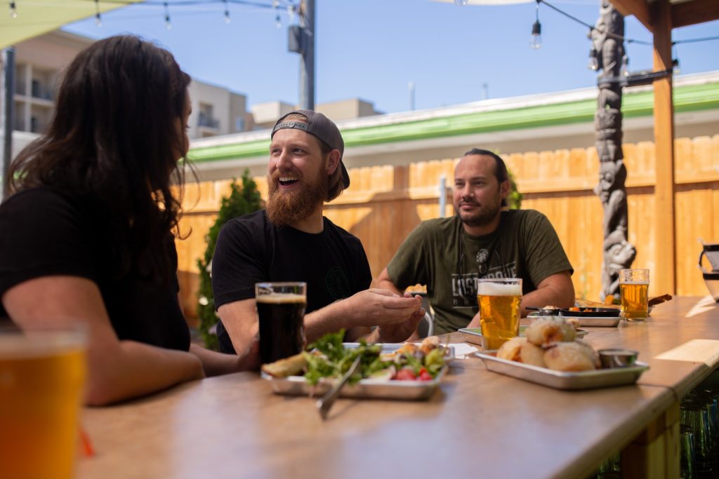 Group of adults enjoying drinks and food on a sunny brewery patio during a trivia night.