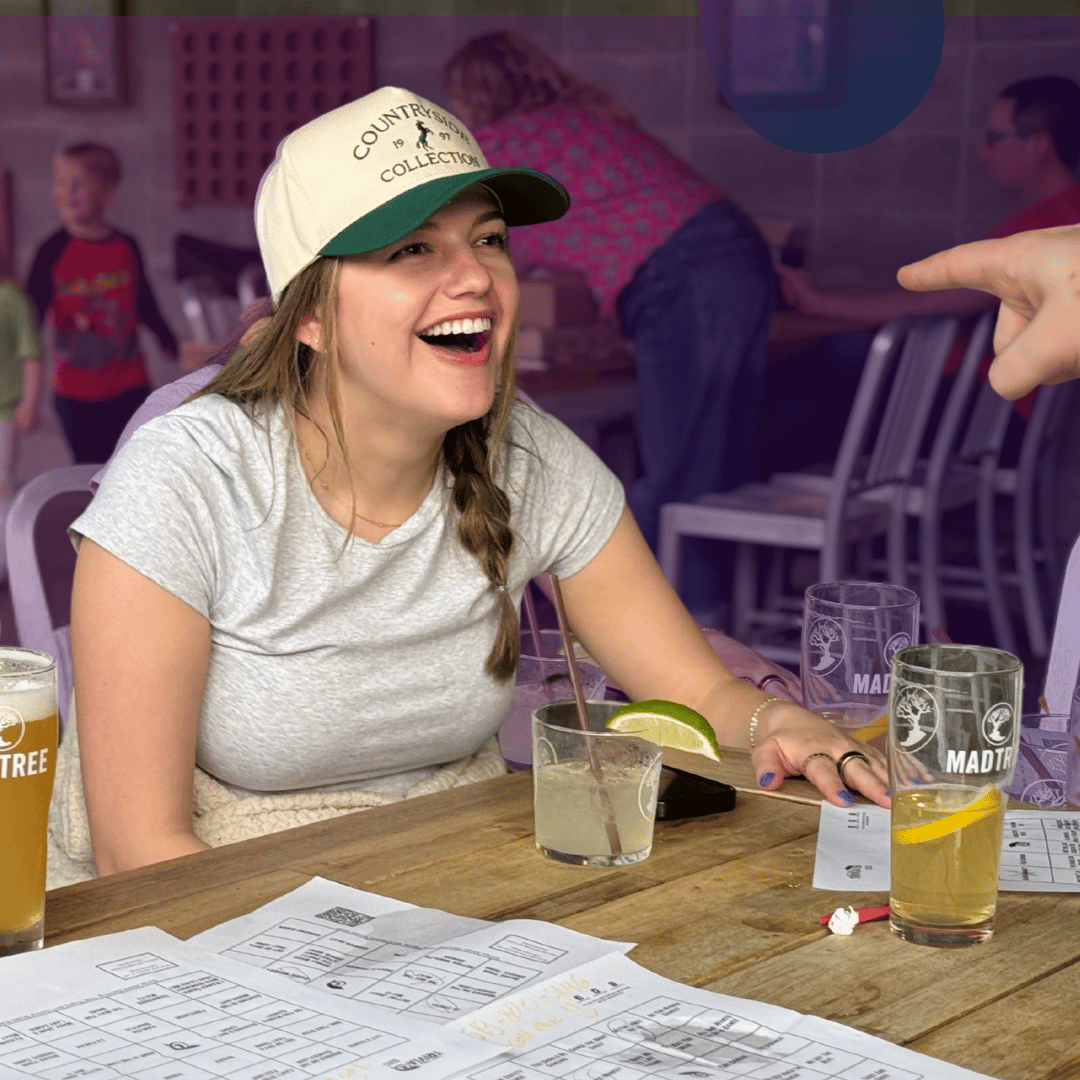 Woman laughing at a bar table during music bingo night with drinks and bingo sheets in front of her
